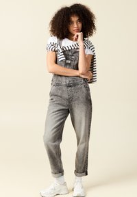 Pantalones de mezclilla grises tipo overol con un fit relajado, tirantes ajustables y bolsillos frontales, combinados con una camiseta a rayas atada a la cintura y zapatillas blancas.