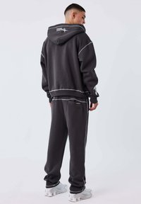 Hombre vistiendo una sudadera con capucha de un gris oscuro y pantalones de chándal a juego con costuras blancas visibles, zapatillas blancas, de pie de espaldas sobre un fondo blanco.