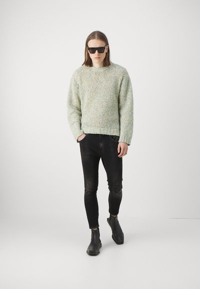 Maglione a maglia texturizzata verde chiaro, pantaloni in denim neri slim-fit e stivali neri alla caviglia. Il modello indossa occhiali da sole quadrati neri. Sfondo minimale.