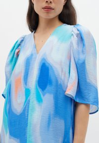 Blauwe jurk met een patroon en pofmouwen, met een V-halslijn. De stof is licht van gewicht in tinten blauw, blauwgroen en koraalaccenten.