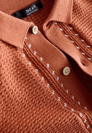 Terrakotowy sweter polo z teksturowaną powierzchnią, kołnierzykiem i dwoma drewnianymi guzikami. Posiada białe ozdobne przeszycia na wysokości klatki piersiowej.
