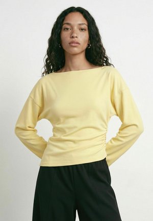 Jeune femme aux longs cheveux bouclés portant un haut à manches longues jaune pâle et un pantalon noir, debout les mains sur les hanches devant un fond uni.