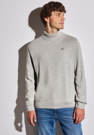 Pull à col roulé gris en tissu doux, avec un col et des poignets côtelés, portant un petit logo sur la poitrine. Associé à un jean bleu.