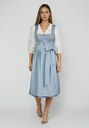 Vrouw draagt een blauwe dirndl met patroon en een effen blauw schort vastgebonden in de taille, een witte kanten blouse en zilveren sandalen met hak, staand.