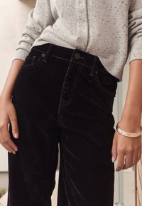 Pantalones de pana negros con bolsillos frontales y cierre de botón, combinados con un cárdigan tejida gris jaspeado. Textura suave y ajuste relajado.