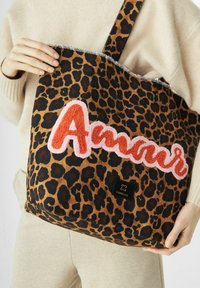 Persoon die een grote tas met luipaardprint vasthoudt, met oranje en roze geborduurde tekst "Amour" en een Codello logo patch.