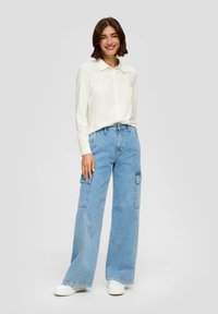 Witte langmouwige button-up shirt, lichtblauwe wijde cargo jeans met voorzakken, en witte sneakers. Eenvoudige, casual outfit.