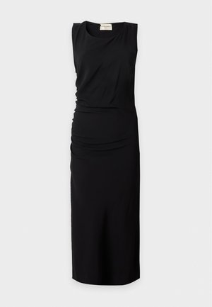 Une femme porte une robe midi noire sans manches et des sandales noires sur un fond clair et uni.