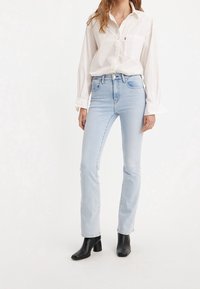 Světle modré džíny rovného střihu vyrobené z denimu, zkombinované s bílou košilí s dlouhým rukávem. Košile má kapsu a zapínání na knoflíky. Černé kotníkové boty dotvářejí vzhled.