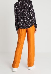 Camicia nera con motivo floreale rosa, abbinata a pantaloni arancioni a pieghe. Scarpe con suola bianca visibili in basso. Colori brillanti e a contrasto.