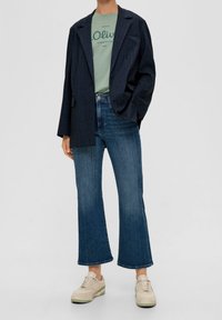 Marinblå blazer, grön grafisk t-shirt och utsvängda blå jeans. Vanliga sneakers fulländar outfiten. Materialen verkar mjuka och avslappnade.