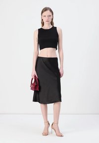 Svart crop topp kombinert med en knelengde svart skjørt som har en subtil glans. Modellen holder en liten rød håndveske og har på seg beige hæler.