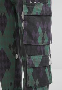 Pantalones cargo de tela negra con un patrón de diamantes en verde y púrpura. Destacan numerosos bolsillos laterales y una textura robusta.