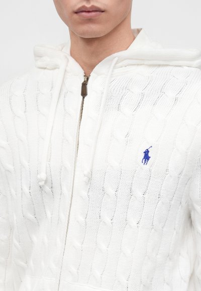 Νεαρό άτομο φοράει ένα λευκό πλεκτό hoodie με φερμουάρ και μπλε κεντημένο λογότυπο Polo στο στήθος και κορδόνια στη κουκούλα.
