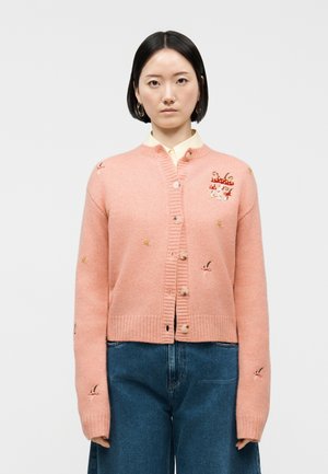 Kvinde med kort sort hår iført en lyserød cardigan med knapper og broderede svampe og fugle, kombineret med blå jeans.