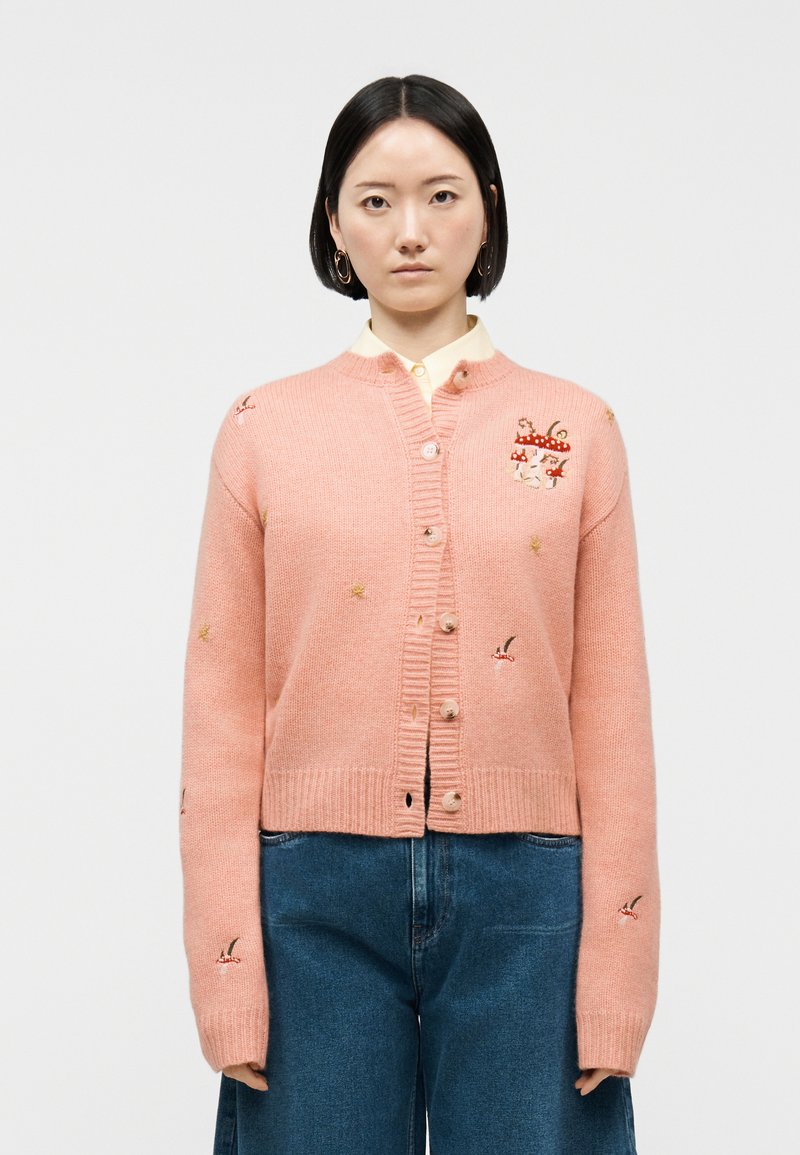 Γυναίκα με κοντά μαύρα μαλλιά που φοράει ροζ ζακέτα με κουμπιά και κεντημένα μανιτάρια και πουλιά, συνδυασμένη με μπλε τζιν.