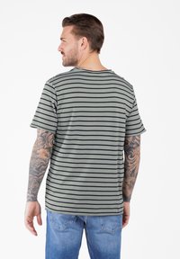 Hombre con brazos tatuados que lleva una camisa de manga corta a rayas grises y negras y jeans azules, girado de espaldas contra un fondo blanco.