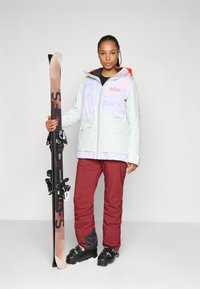 Abbigliamento da sci con giacca verde menta chiaro dal design multicolore, dettagli rossi e pantaloni bordeaux. Attrezzatura da sci tenuta in una mano, stivali neri.