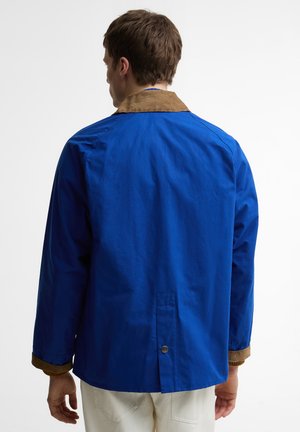 Mees, kes kannab sinist jopet pruunide krae ja mansettidega, vaadatuna tagantpoolt tavalise tausta ees.