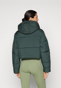 Veste courte en matelassé verte avec une capuche, comportant des sections matelassées et une texture lisse. Associée à des leggings taille haute vert clair.