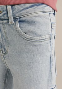 Světlé modré denimové džíny zblízka, s přední kapsou, jemným opotřebením a hladkou texturou s kontrastními švy.