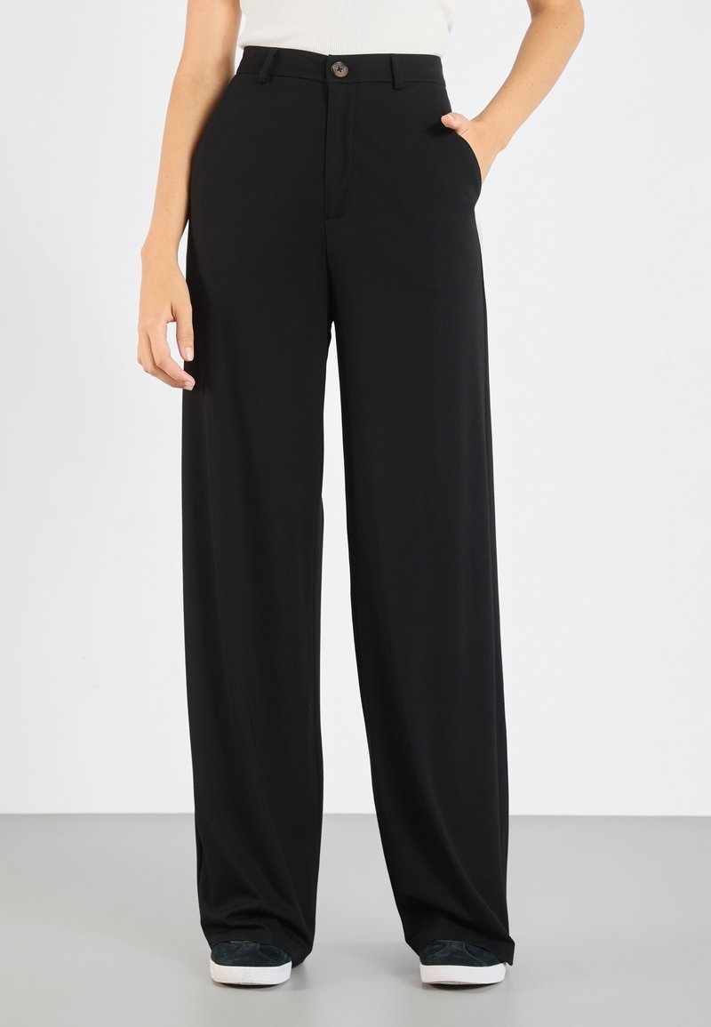 Pantaloni neri a gamba larga realizzati in tessuto morbido, con chiusura a bottone, tasche laterali e una vestibilità comoda, indossati con sneakers scure.