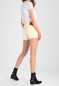 Pantalones cortos de mezclilla amarillo claro con dobladillos deshilachados, combinados con una camiseta blanca y botines negros hasta el tobillo. Presenta un parche de cuero con el logotipo en la parte trasera.