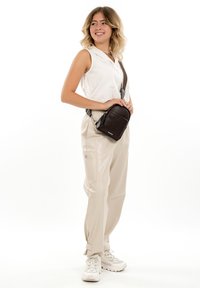 Smiling woman in sleeveless white blouse, beige cargo pants, and white sneakers holding a black crossbody bag with wide strap.