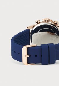Cinturino per orologio in silicone blu navy con fibbia in oro rosa. Il retro dell'orologio presenta una finitura argentata con dettagli incisi e superficie testurizzata.