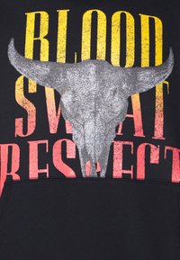 Sudadera negra con un estampado gráfico de un cráneo de toro plateado. El texto dice "SANGRE SUDOR RESPETO" en tonos amarillos, rojos y rosas, con un acabado envejecido.