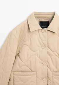 Beżowa pikowana kurtka o falistej fakturze, z kołnierzykiem koszulowym, guzikami z przodu oraz dwoma bocznymi kieszeniami. Metka widoczna wewnątrz kołnierza.