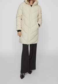 Woman wearing a beige quilted long coat with hood, black wide-leg pants, and black pointed shoes standing against a plain background.