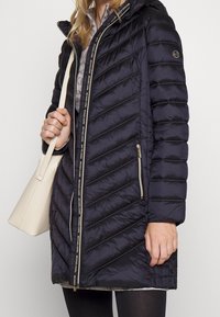 Abrigo de plumas navy con cierre de cremallera y patrón de acolchado diagonal. Presenta una capucha y un detalle de logo a lo largo de la cremallera. Estilizado con un bolso ligero.