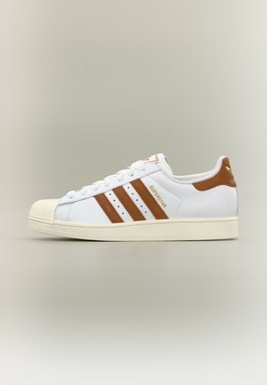 Witte Adidas Superstar sneaker met bruine strepen en hieltab, crèmekleurige zool en gestructureerde neuskap, getoond in zijaanzicht op een neutrale achtergrond.