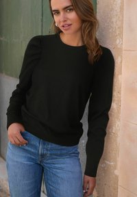 Maglione nero con maniche a sbuffo, polsini e orlo a coste, abbinato a jeans blu chiaro. Tessuto morbido in maglia.