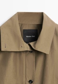 Khakibrauner Mantel mit Kragen und schwarzen Knöpfen sowie Massimo Dutti Etikett im Kragen, vor weißem Hintergrund präsentiert.