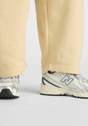 Beige Hosen, die teilweise weiße und silberne New Balance Sneakers bedecken, stehen auf einer grauen Oberfläche.