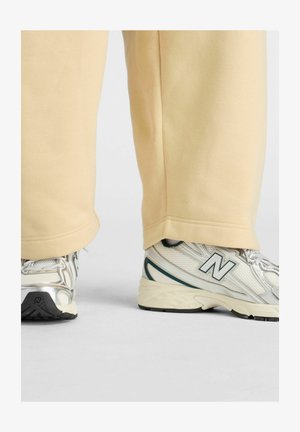 Beige broek die gedeeltelijk witte en zilveren New Balance sneakers bedekt, staand op een grijze ondergrond.