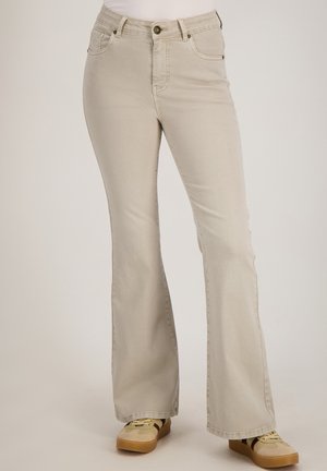 Licht beige bootcut jeans gedragen met beige en bruine sneakers, weergegeven van taille tot voeten tegen een effen achtergrond.