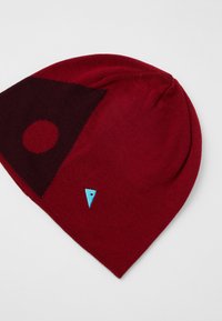 Gorra de punto roja con un diseño geométrico de color marrón oscuro que incluye un círculo y acentos triangulares, junto con un pequeño emblema azul.