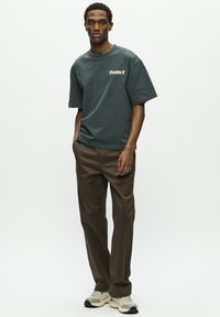Graues Baumwoll-T-Shirt mit "Double V"-Logo auf der Brust, kombiniert mit braunen Hosen und hellgrauen Sneakers mit dunklen Akzenten.