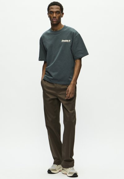 T-shirt en coton gris avec le logo "Double V" sur la poitrine, associé à un pantalon marron et des baskets gris clair avec des accents foncés.