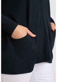Cardigan a maglia navy con polsini a coste, dotato di due tasche frontali, vestibilità comoda e texture liscia. Sfondo neutro bianco.
