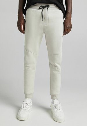Homme portant un pantalon de jogging beige clair avec un cordon noir et des baskets blanches, debout devant un fond gris uni.