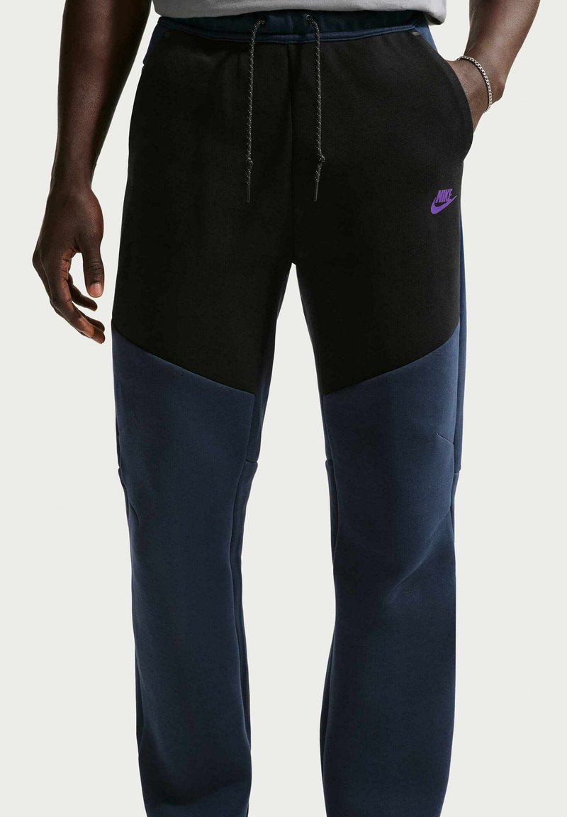 Pantalones deportivos Nike con diseño de bloques de color negro y azul marino, cintura elástica y bolsillos laterales visibles con un detalle de logo púrpura.