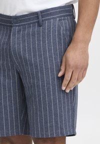 Modré pruhované šortky vyrobené z jemnej látky, s gombíkovým zapínaním, prednými skladmi a uvoľneným strihom. Pruhy sú svetlomodré na tmavom pozadí.