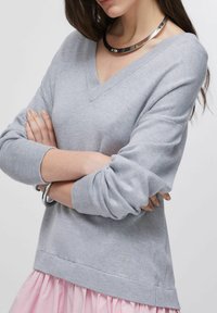 Pull gris à col en V en maille douce avec des poignets côtelés, présentant un petit détail orné à l'ourlet, porté sur une jupe rose à volants.