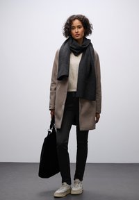 Cappotto in lana beige sopra un maglione crema, jeans neri, sciarpa grigia e sneakers scure, mentre sorregge una borsa a tracolla nera. Design minimalista, colori neutri.