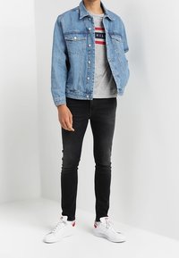 Chaqueta de mezclilla azul claro sobre una camiseta gris con rayas rojas y azul marino, combinada con jeans negros ajustados y zapatillas blancas con detalles en rojo.