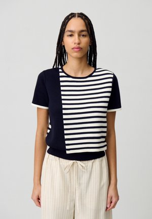 Suéter de punto de manga corta con un frente a rayas en azul marino y blanco, parte trasera sólida en azul marino y un dobladillo acanalado. Combinado con pantalones ligeros a rayas.